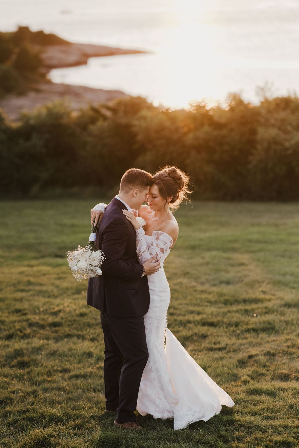 Newlyweds embrace on hilltop overlooking water during sunset.