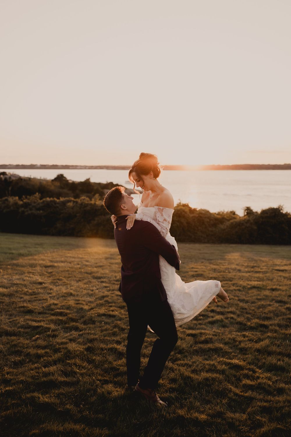 Groom lifts bride in white dress against sunset waterfront view.