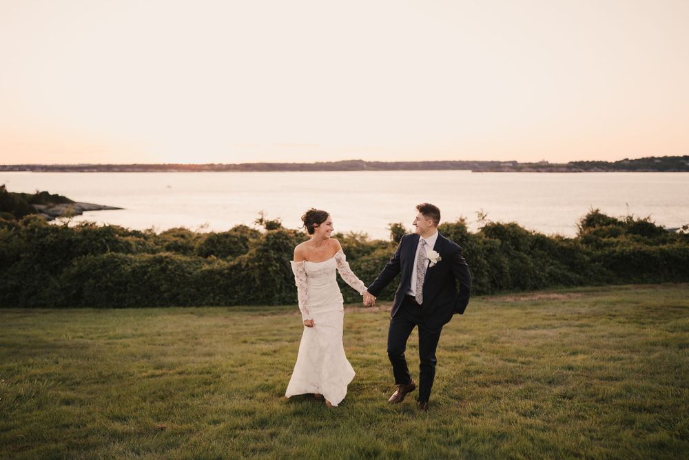 Newlyweds dance together on grassy hilltop with water view at sunset.