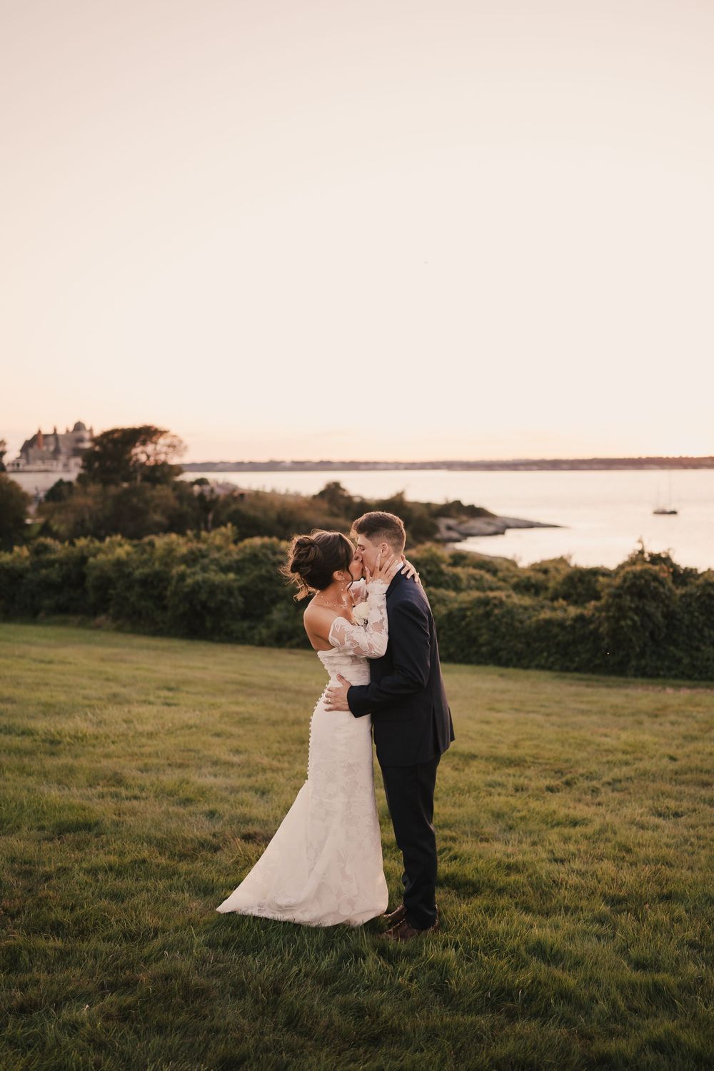 A romantic wedding couple embraces on a grassy hill overlooking the ocean at sunset.