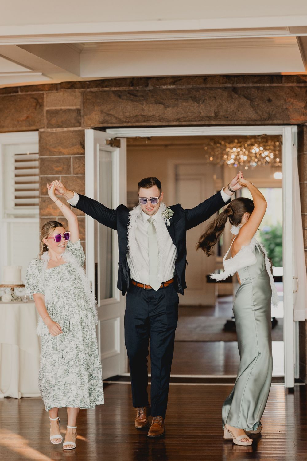 Wedding party members dance joyfully in sunglasses at an indoor reception venue.