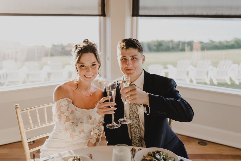 Newlyweds toast with champagne glasses at their reception table by large windows.