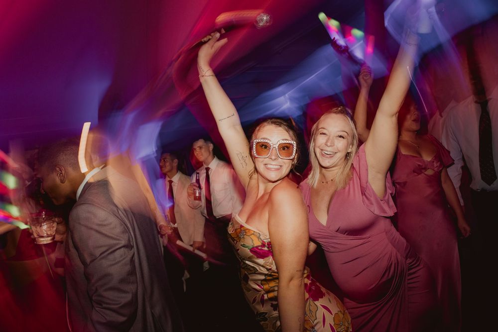 Party guests dancing energetically under vibrant pink and purple lighting at an event.