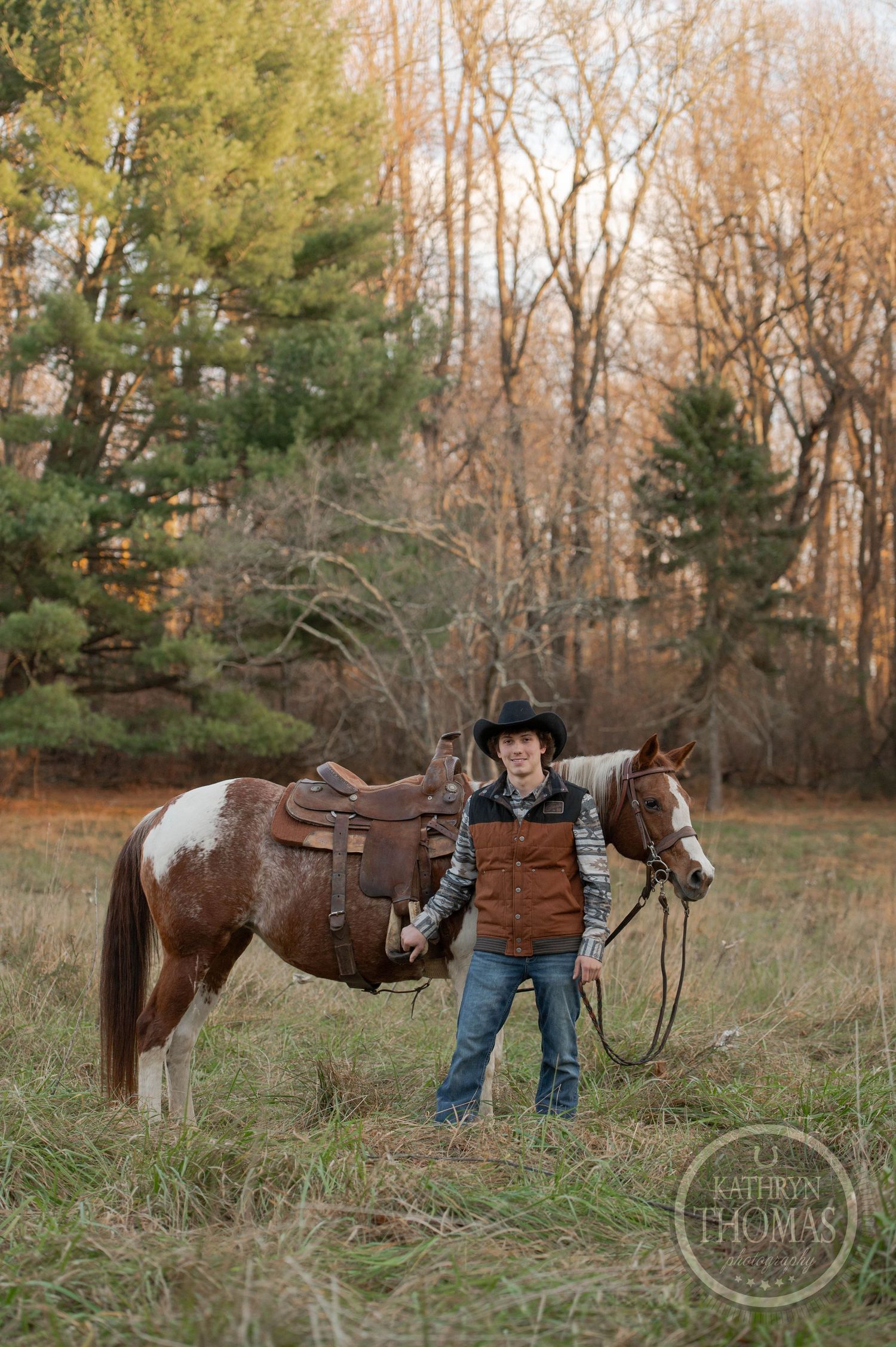 A paint horse with leather saddle stands with a rider in brown vest and cowboy hat in an autumn field.