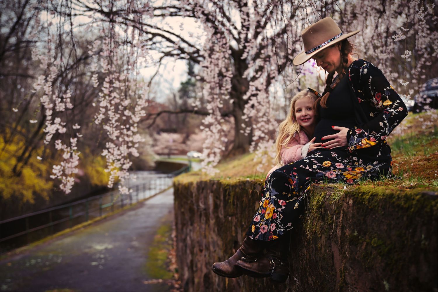 Maternity photo of mother and child sitting on stone wall beneath blooming Cherry trees near pathway.