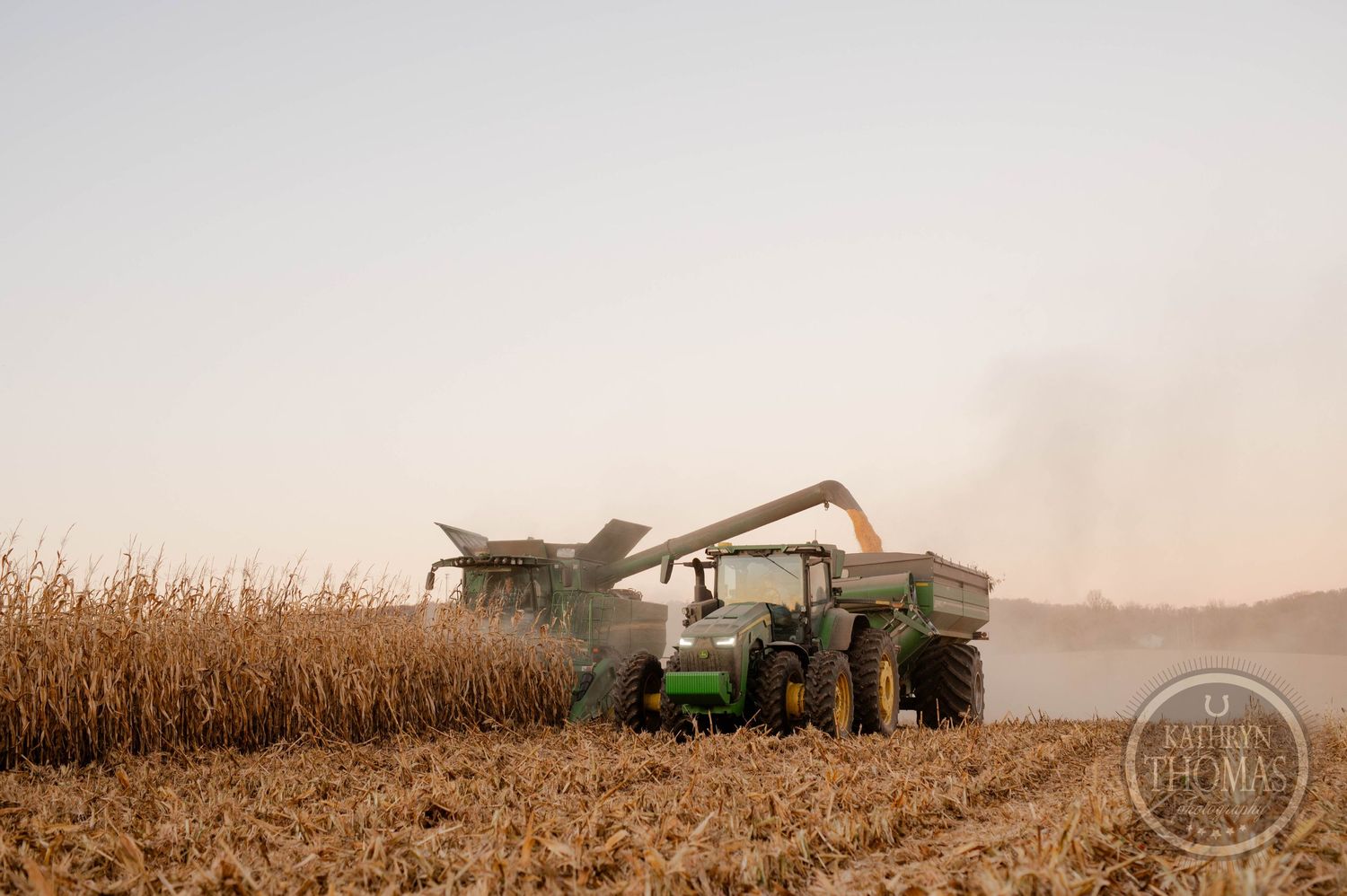 Large combine harvester and tractor working together to harvest a cornfield at sunset.
