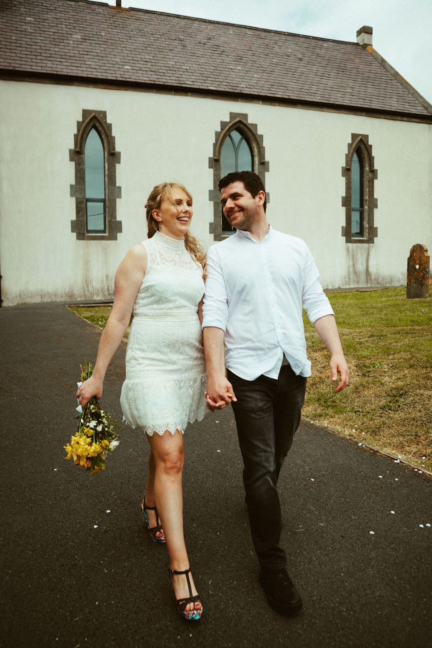A loving couple walks down a paved path outside a church, wearing wedding attire and holding a yellow bouquet.