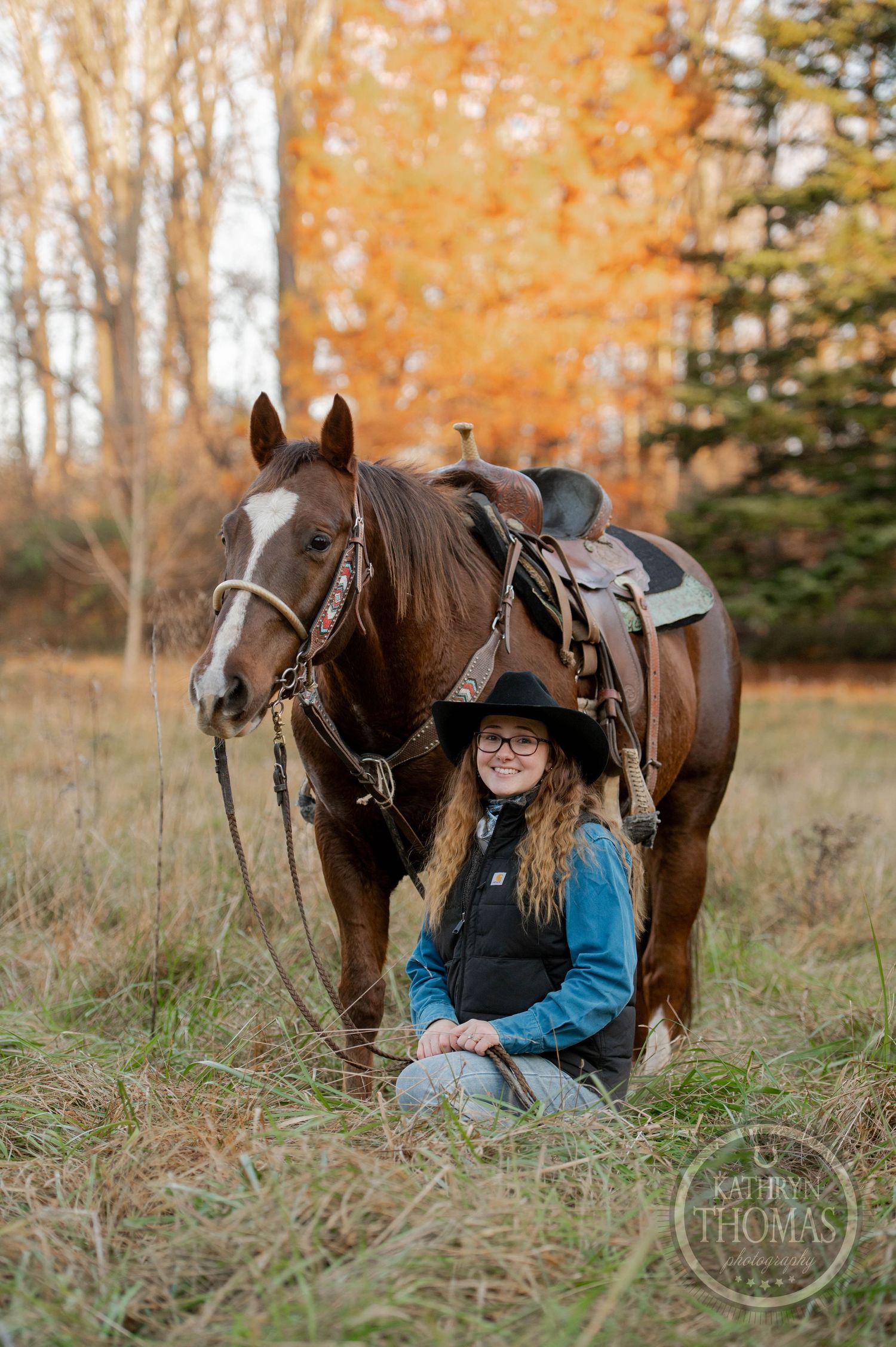 A brown horse with leather saddle stands next to a seated figure in a western hat and blue jacket during autumn.