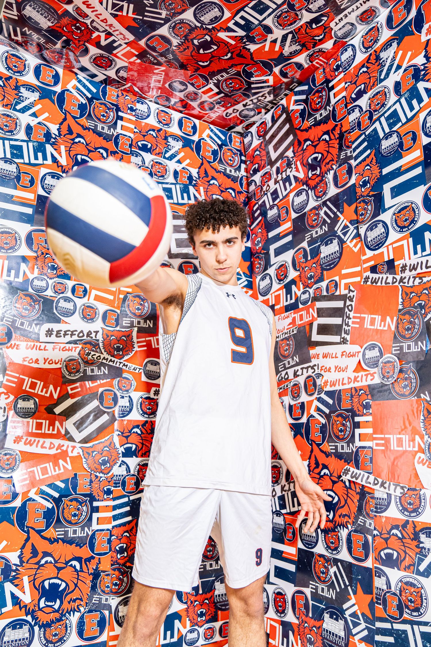 Athlete in white uniform poses with volleyball against orange and blue patterned backdrop with team logos.