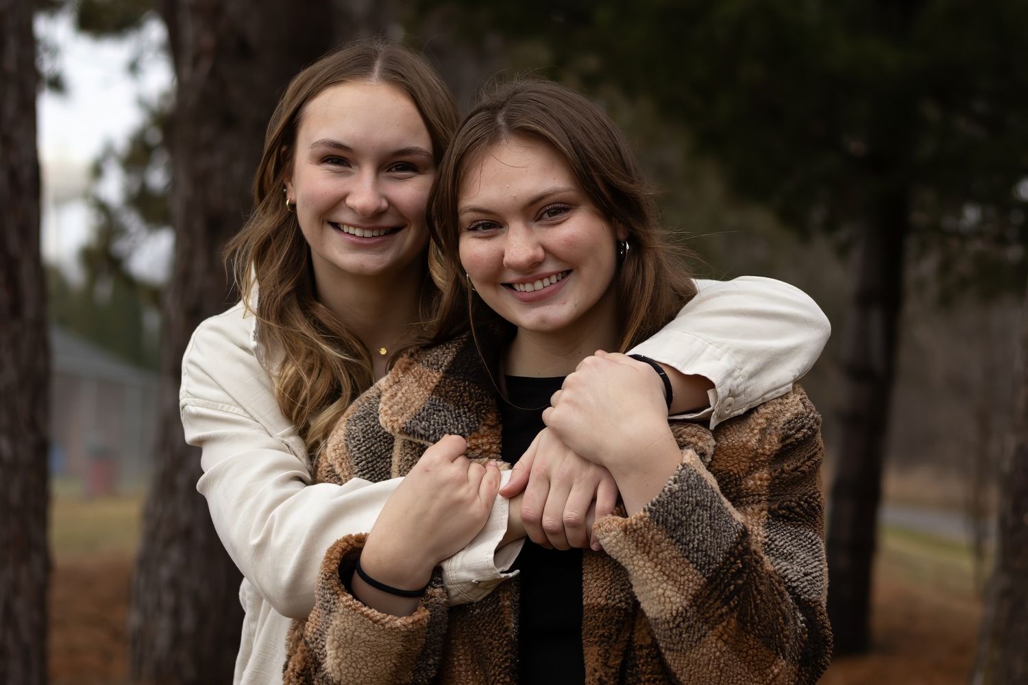Two young women in cream-colored sweaters embrace and smile together in a wooded outdoor setting.