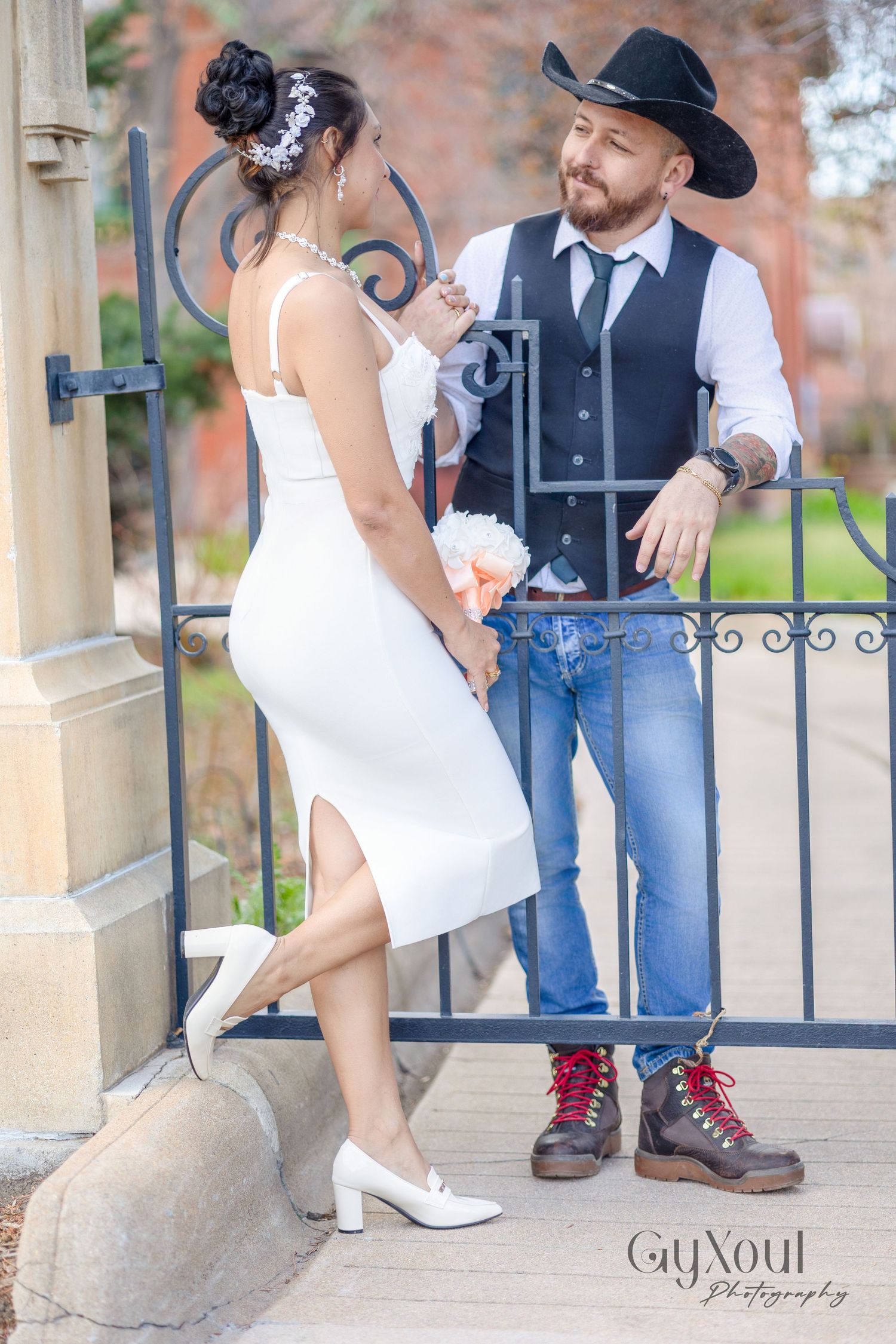 A couple in wedding attire embrace by an ornate iron gate, with the bride in white and groom in western cowboy style.