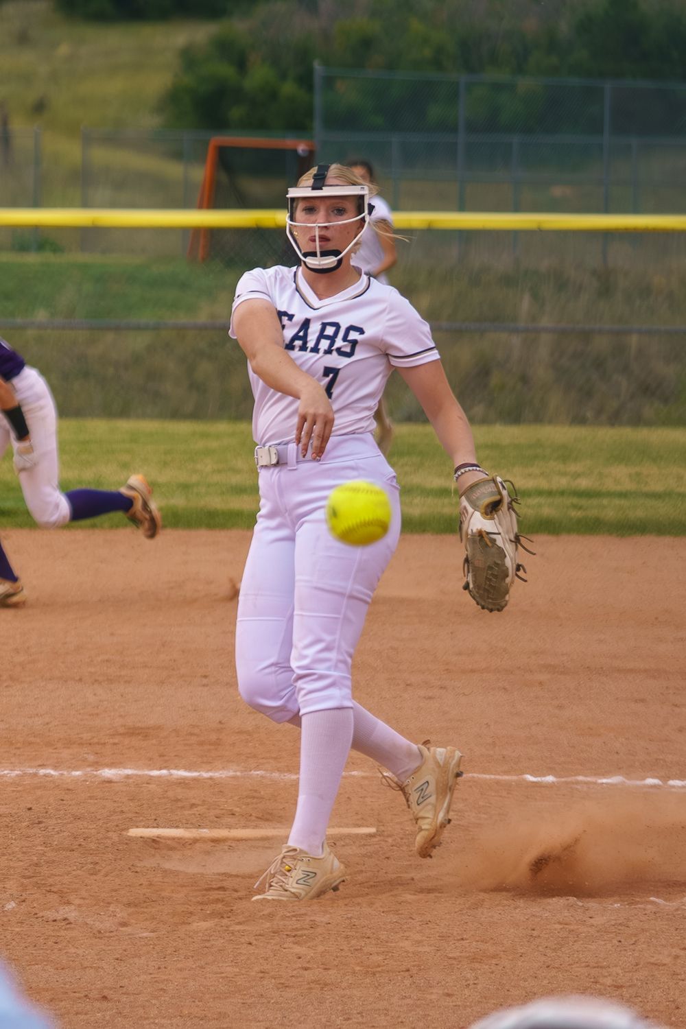 A softball pitcher in white uniform releasing a yellow ball in a sequence of motion on a dirt field.