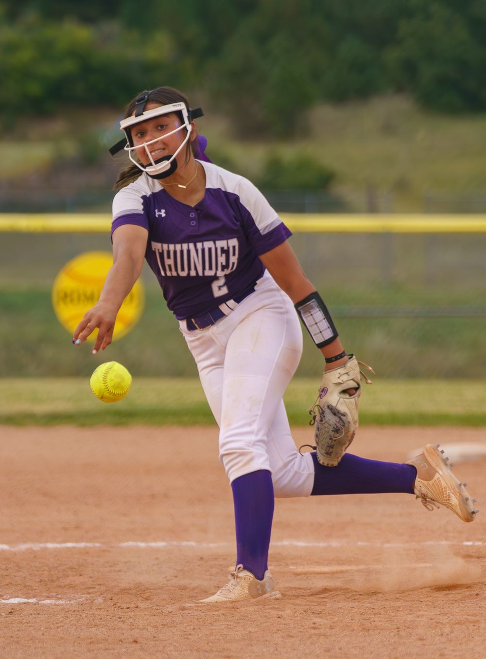 A softball player in purple uniform pitching a yellow ball in a dynamic action sequence.