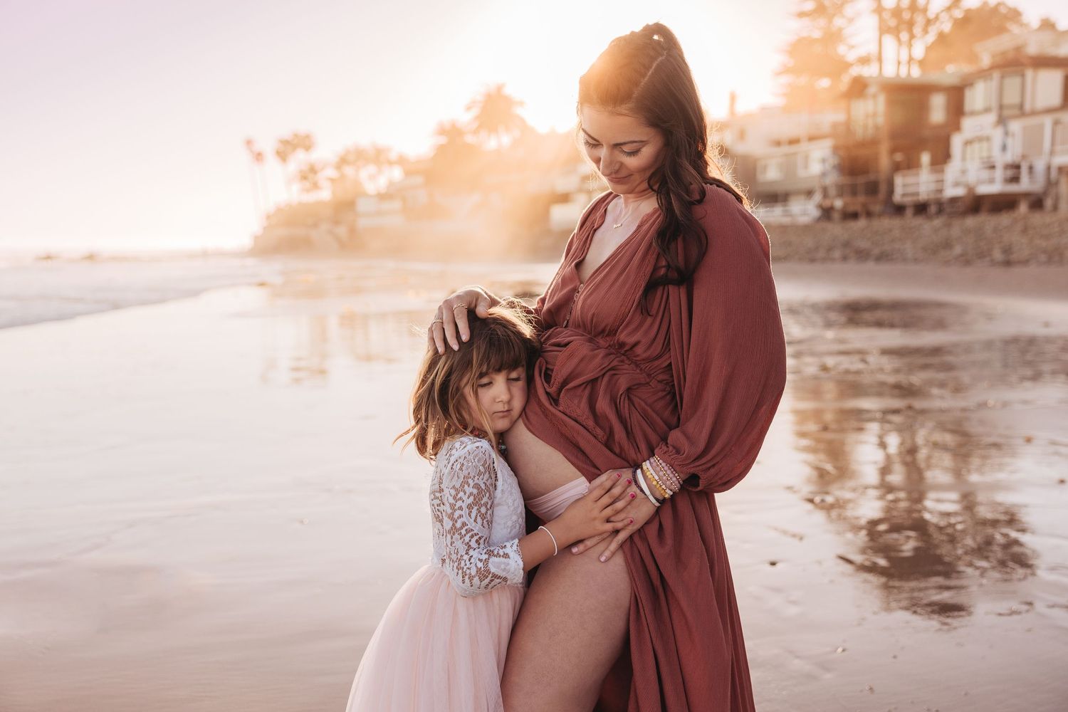 A pregnant woman in a burgundy dress stands with a young child in a white dress on a misty beach at sunset.