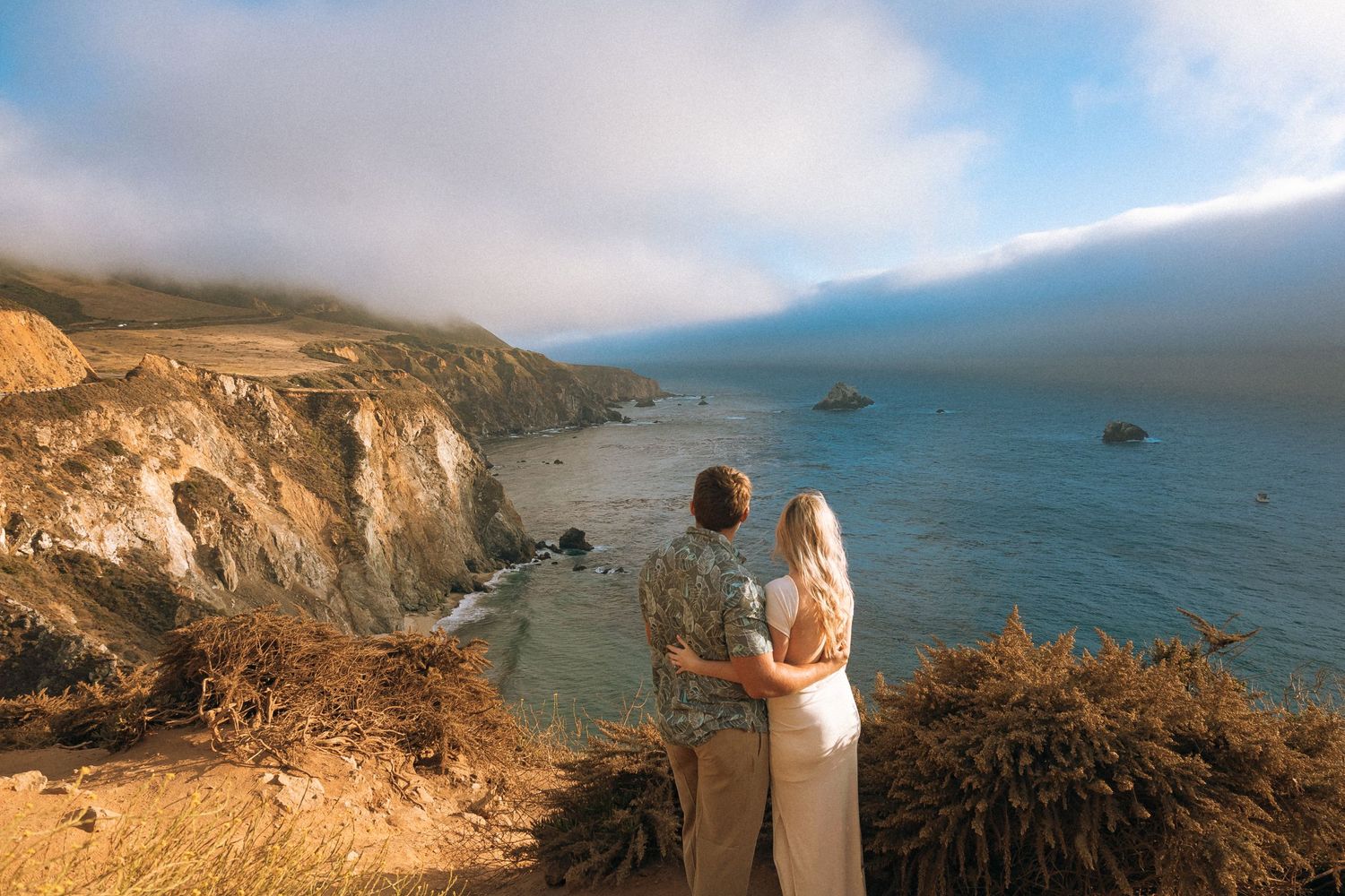 Couple embraces while watching sunset over dramatic coastal cliffs and ocean views at Big Sur, California.