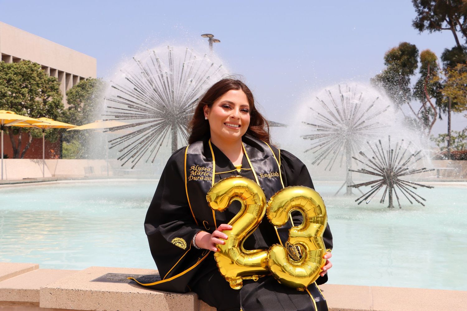 Graduate with gold number balloons celebrating by campus fountain with metal sculptures.