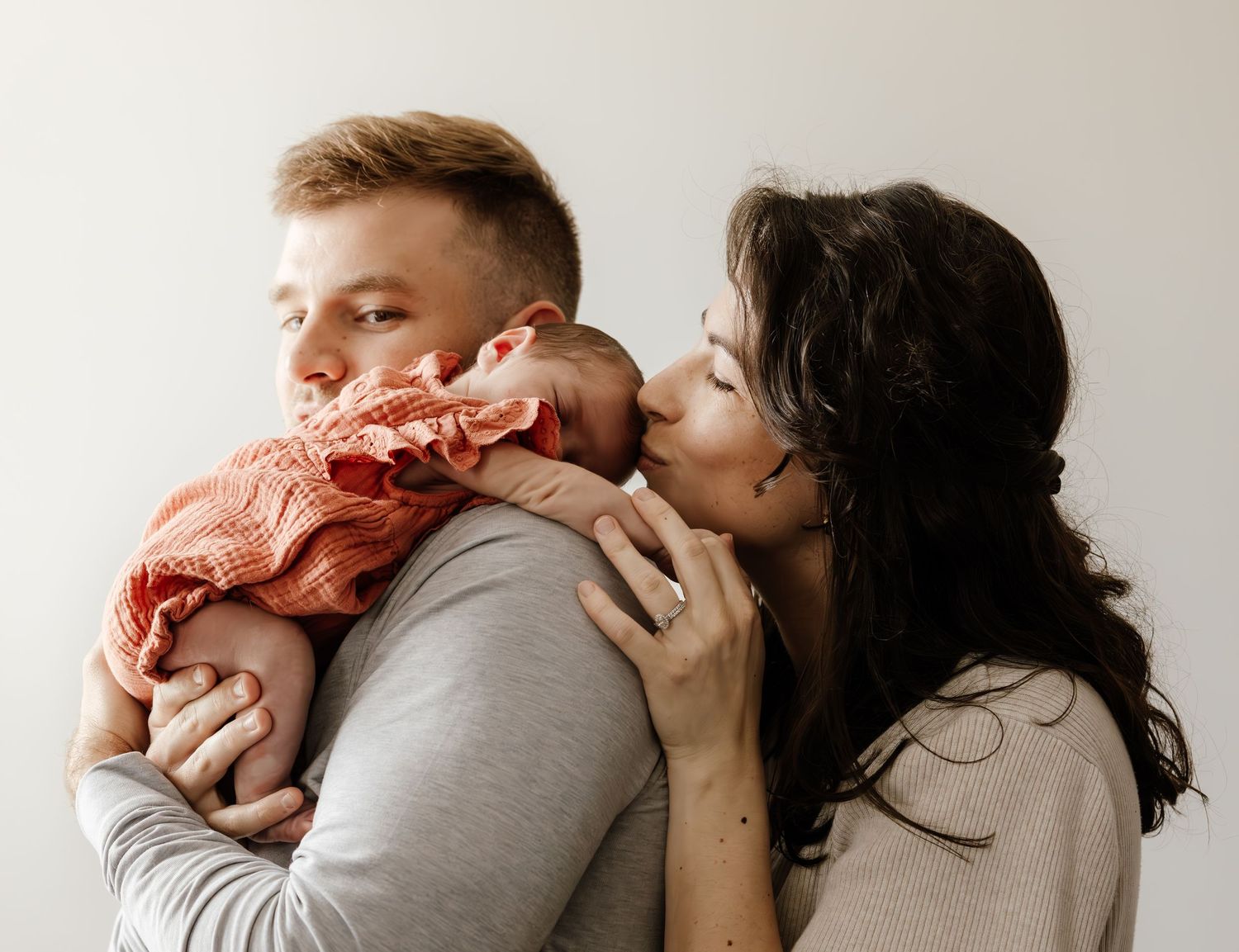A couple in a tender moment with their baby wearing a coral-colored outfit against a neutral background.