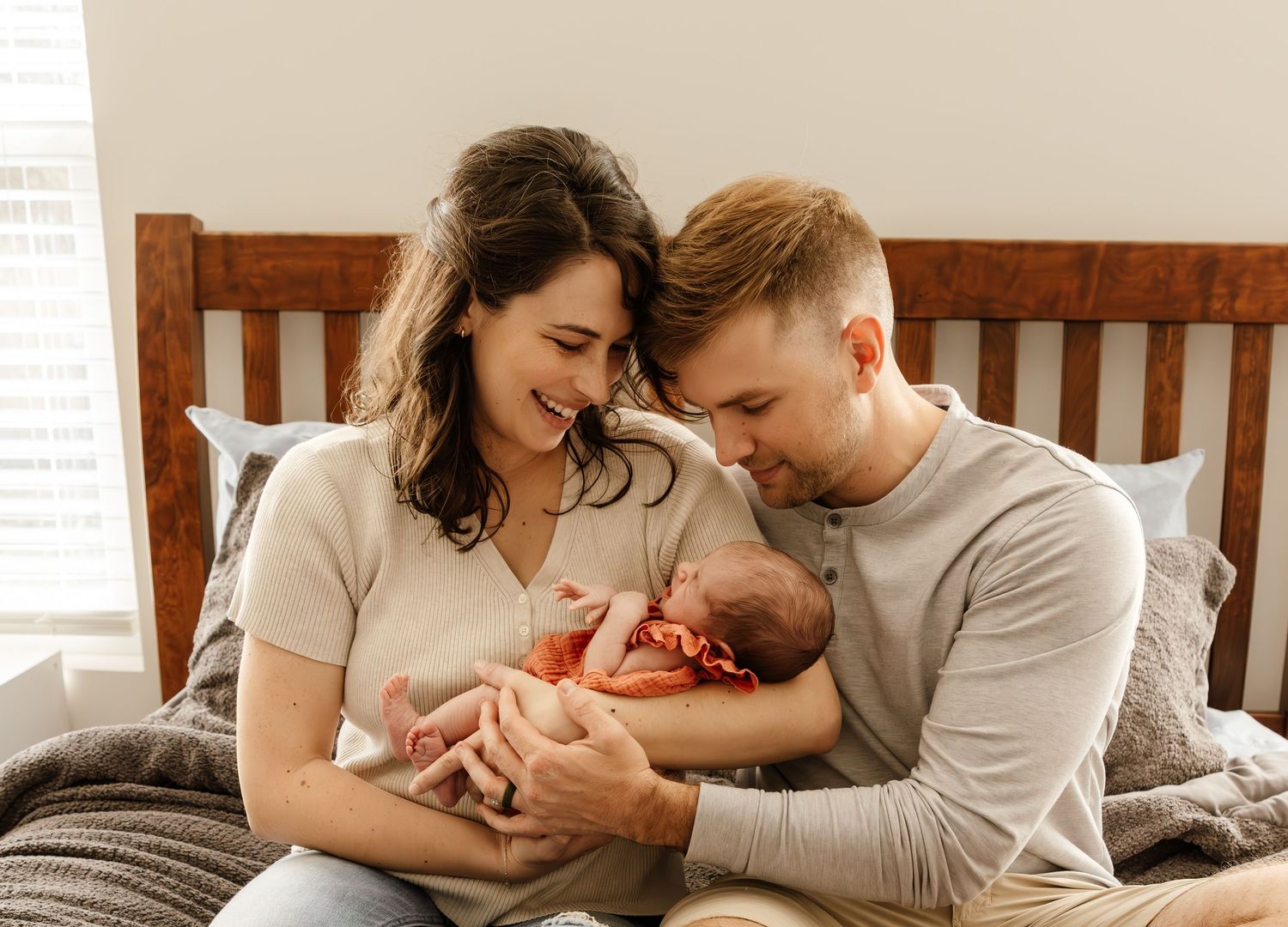 Two parents sitting on bed share tender moment with their newborn baby in cream-colored bedroom setting.
