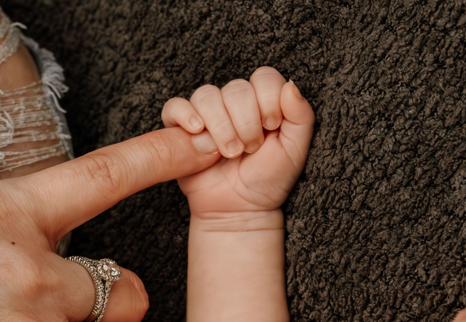 A tiny baby hand grasps an adult finger with a diamond ring against a dark grey textured background.