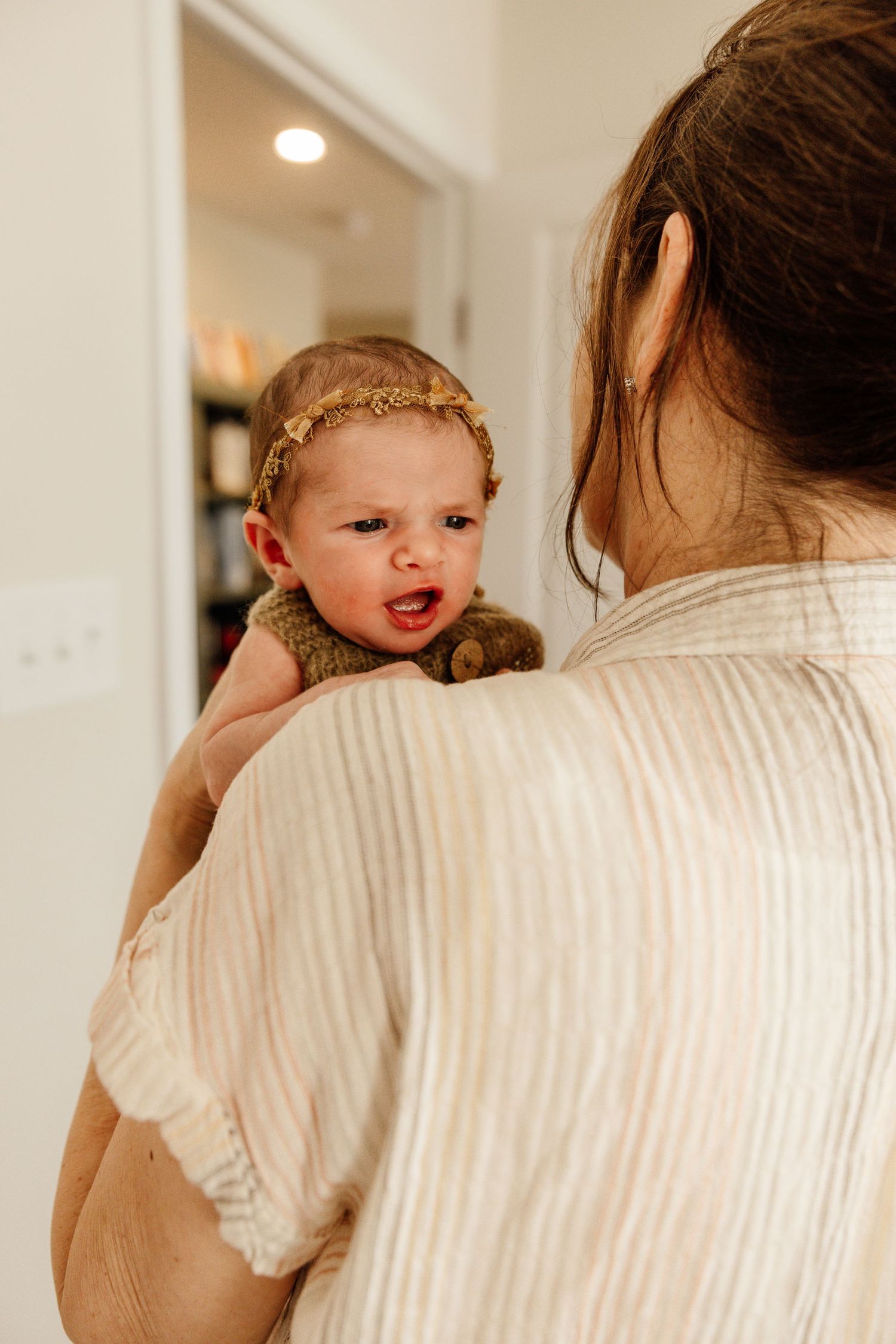 Baby in a brown onesie smiles over the shoulder of someone wearing a white ribbed sweater in warm indoor lighting.