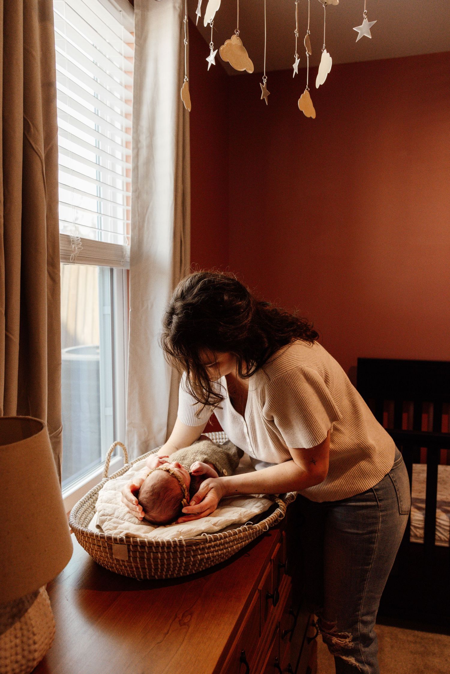 A parent tends to a newborn baby on a woven basket bed by a window with hanging star decorations in a cozy nursery.