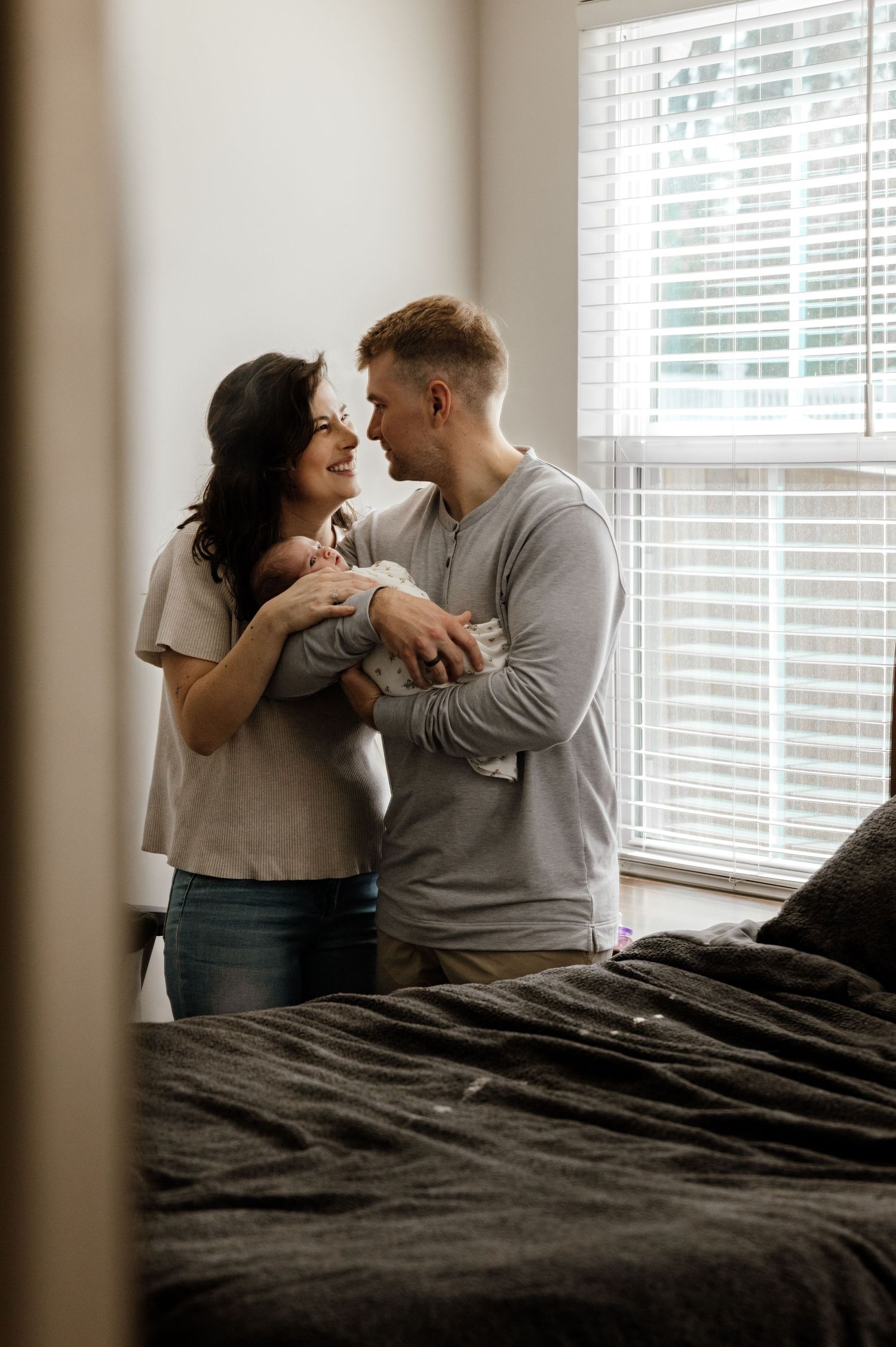 A tender moment between a couple holding coffee cups while standing near a window with venetian blinds in a bedroom.