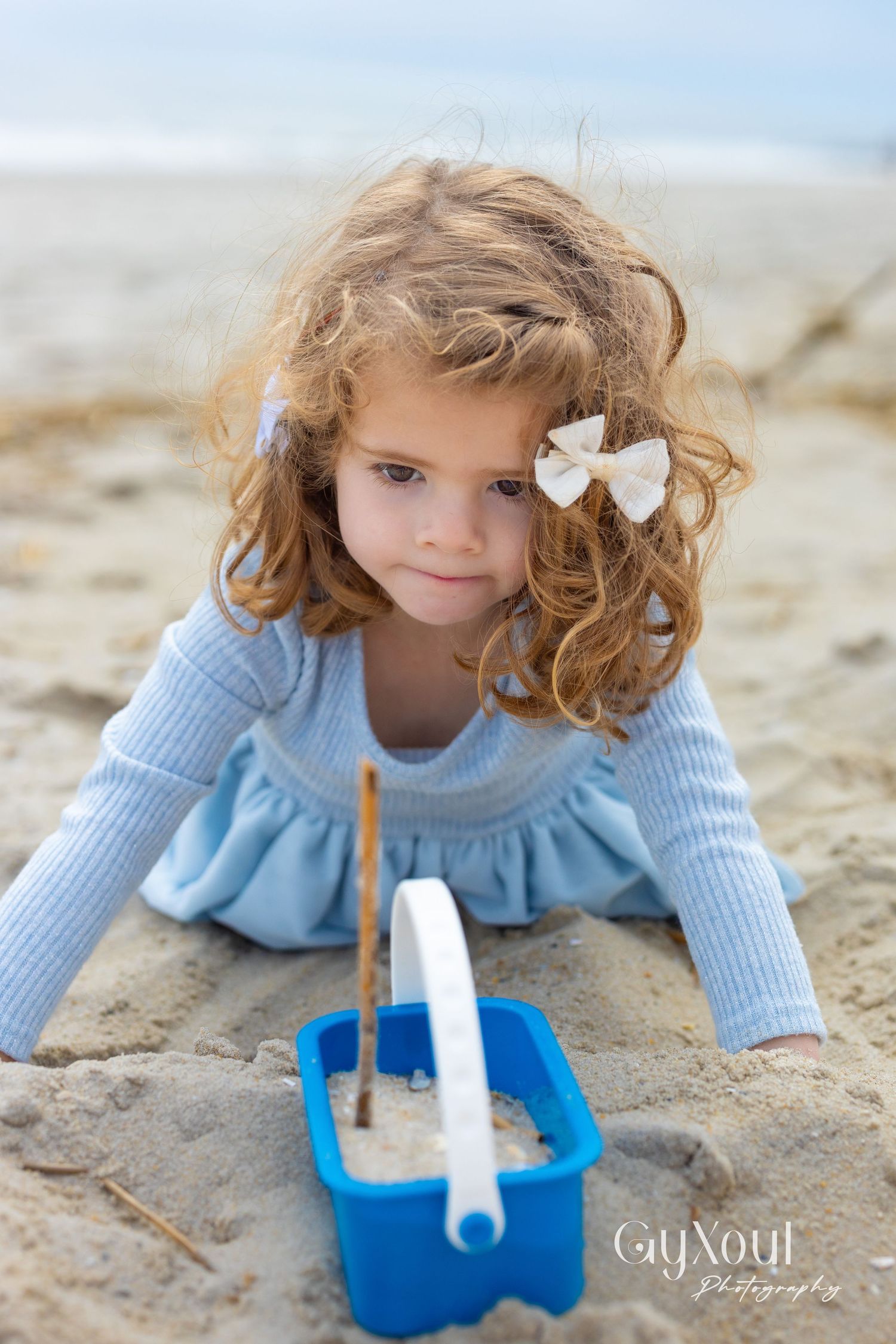 A child in a light blue sweater plays in the sand with a toy boat at the beach on a cloudy day.