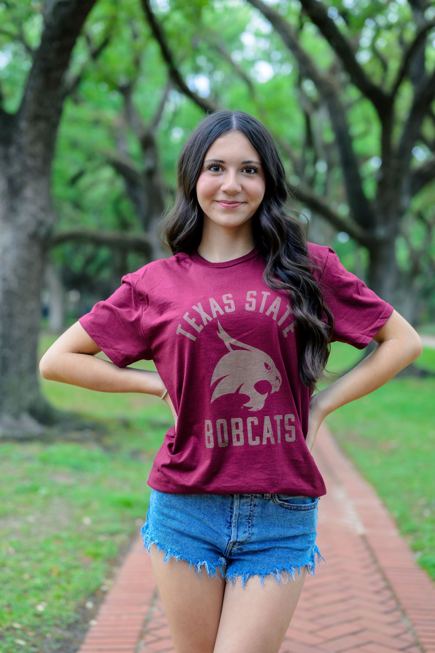 A Texas State Bobcats fan poses on a brick path wearing a maroon team t-shirt and denim shorts among green trees.
