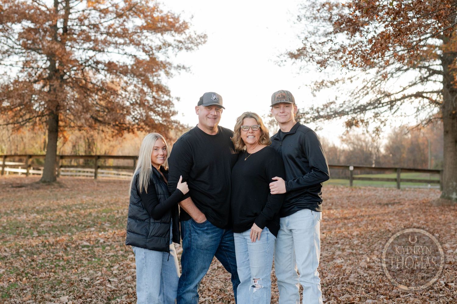 Family wearing matching black tops and jeans pose together in an autumn park setting.
