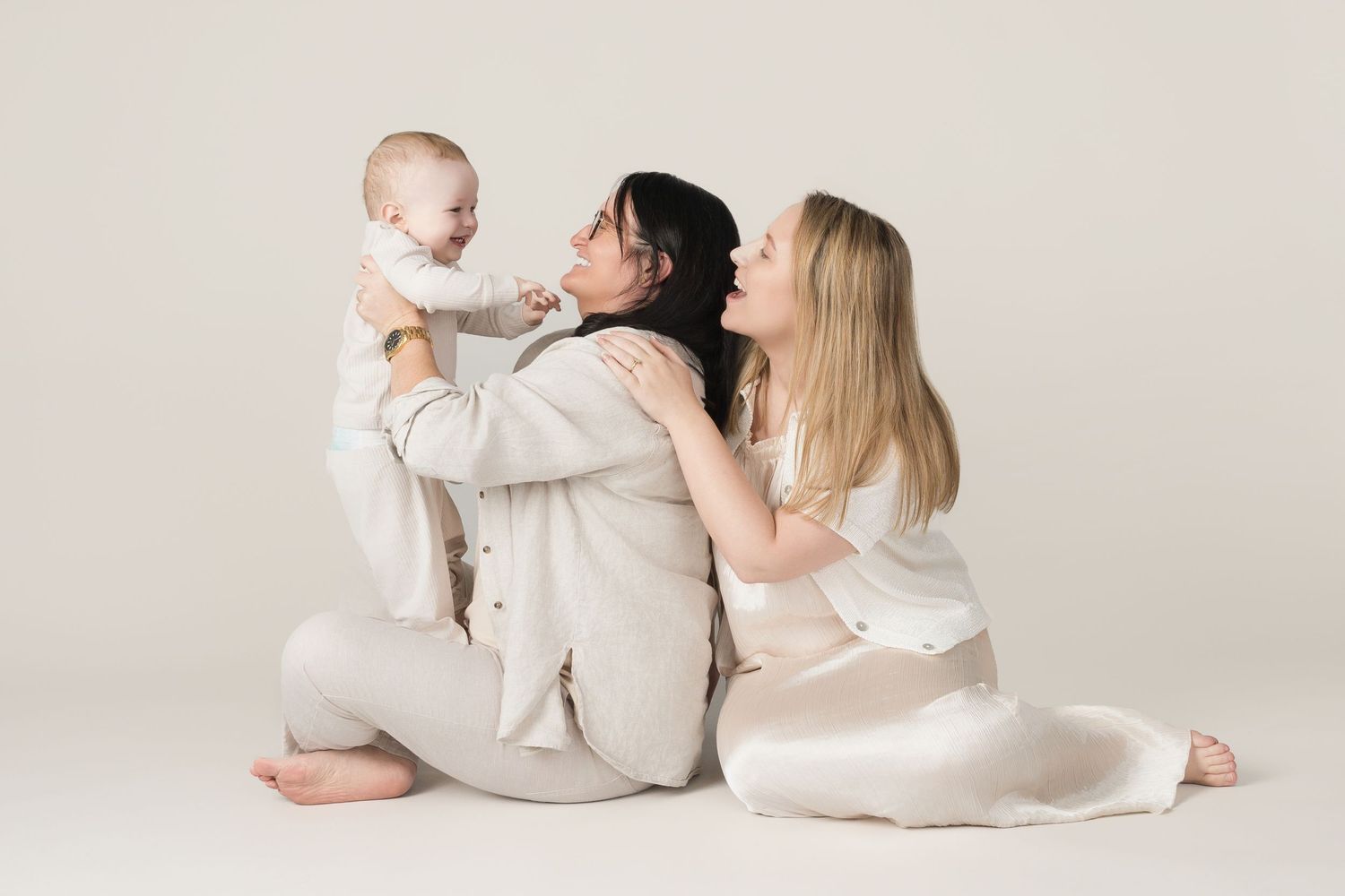 Family wearing neutral-toned outfits sits together on light beige background in studio setting.