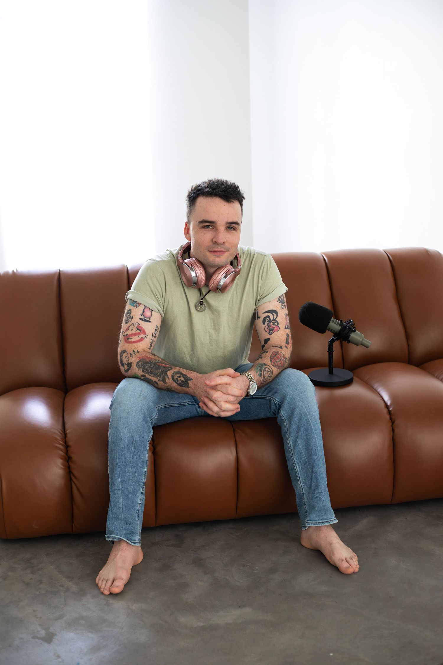 Podcast host with tattoos relaxes barefoot on brown leather couch with microphone setup.