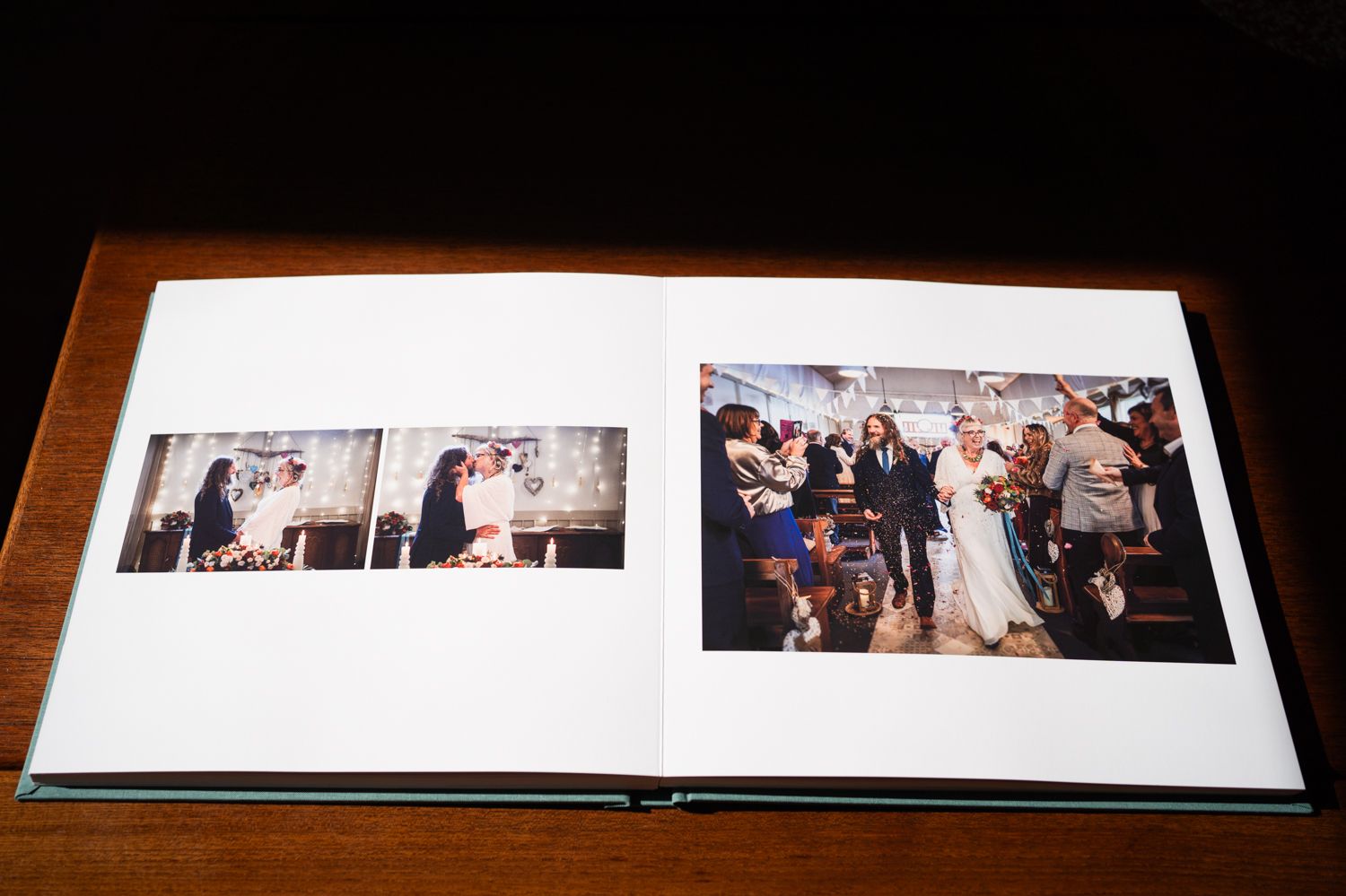 A wedding photo album lies open on a wooden table showing candid reception photographs spread across white pages.