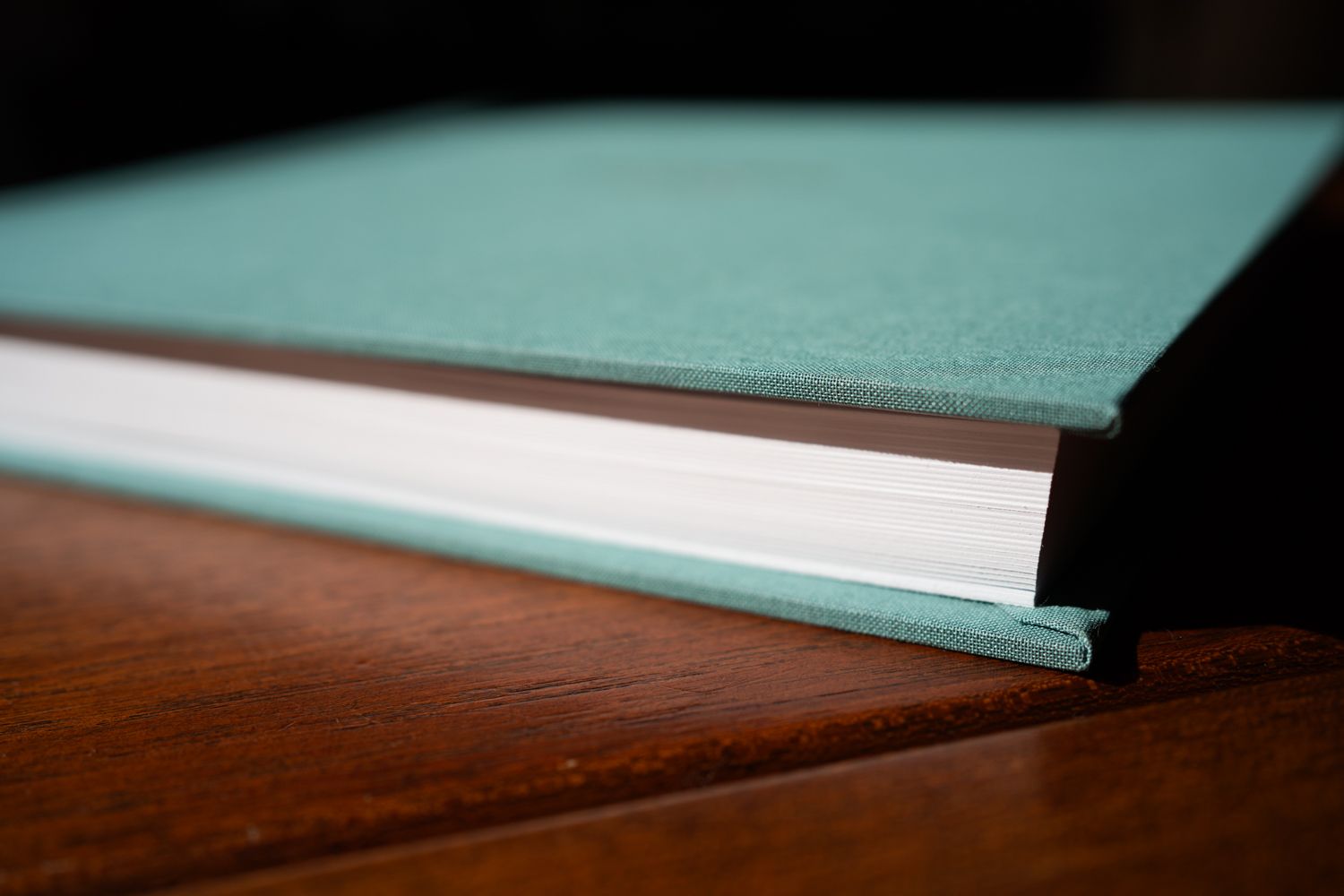 A hardbound book with a teal-colored cover lies on a dark wooden surface, showing its profile view and binding.