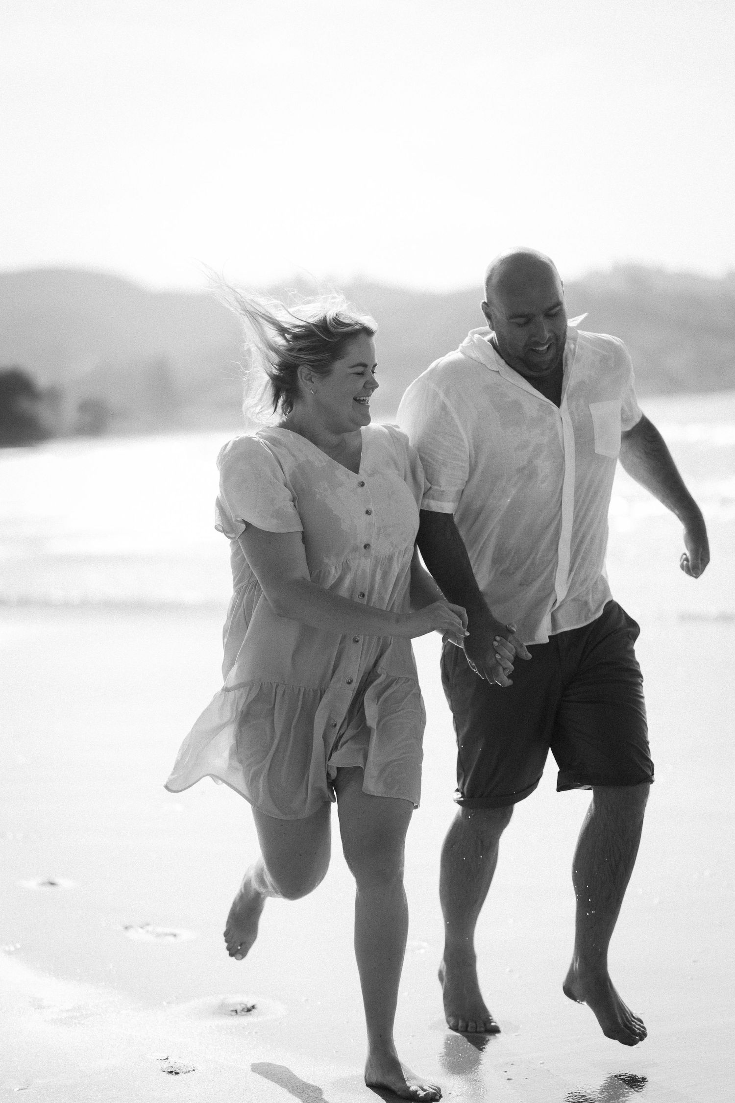 A couple runs joyfully together hand-in-hand along a sunlit beach in a candid black and white photograph.