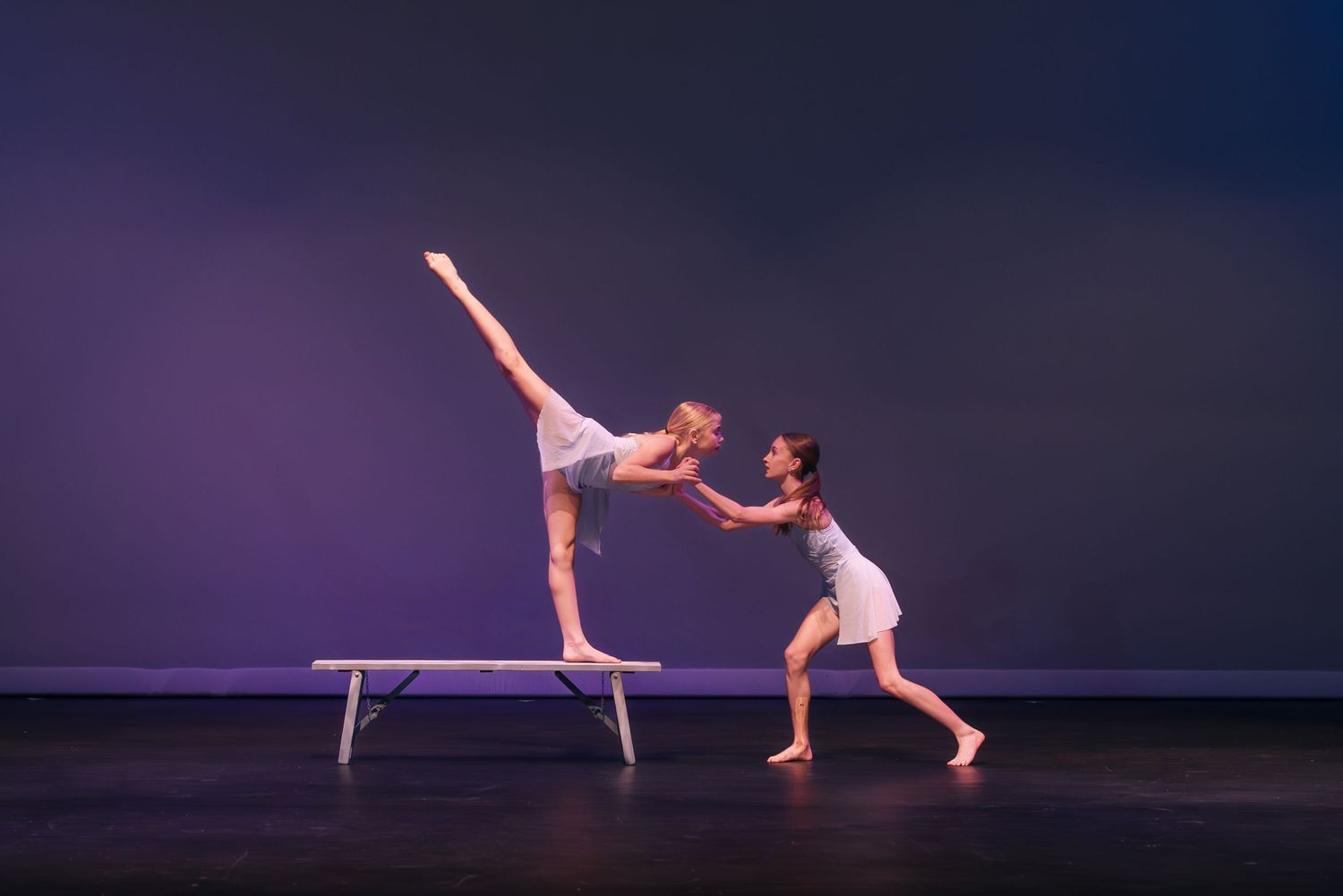 Two ballet dancers perform a dramatic duet with one balanced on a bench while the other supports from below on stage.