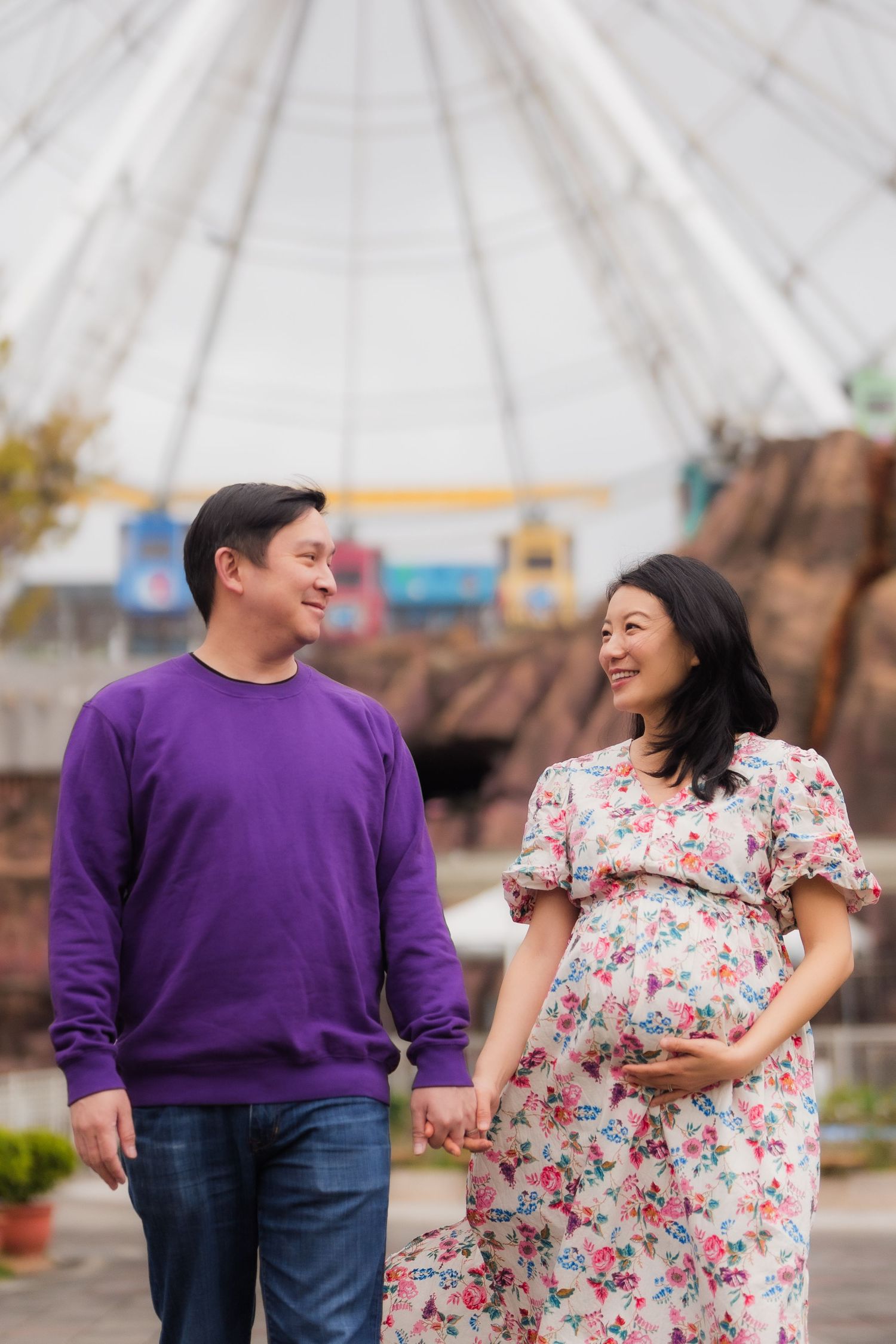 Expectant couple holding hands and smiling while walking outdoors, with woman wearing floral dress and man in purple sweater.