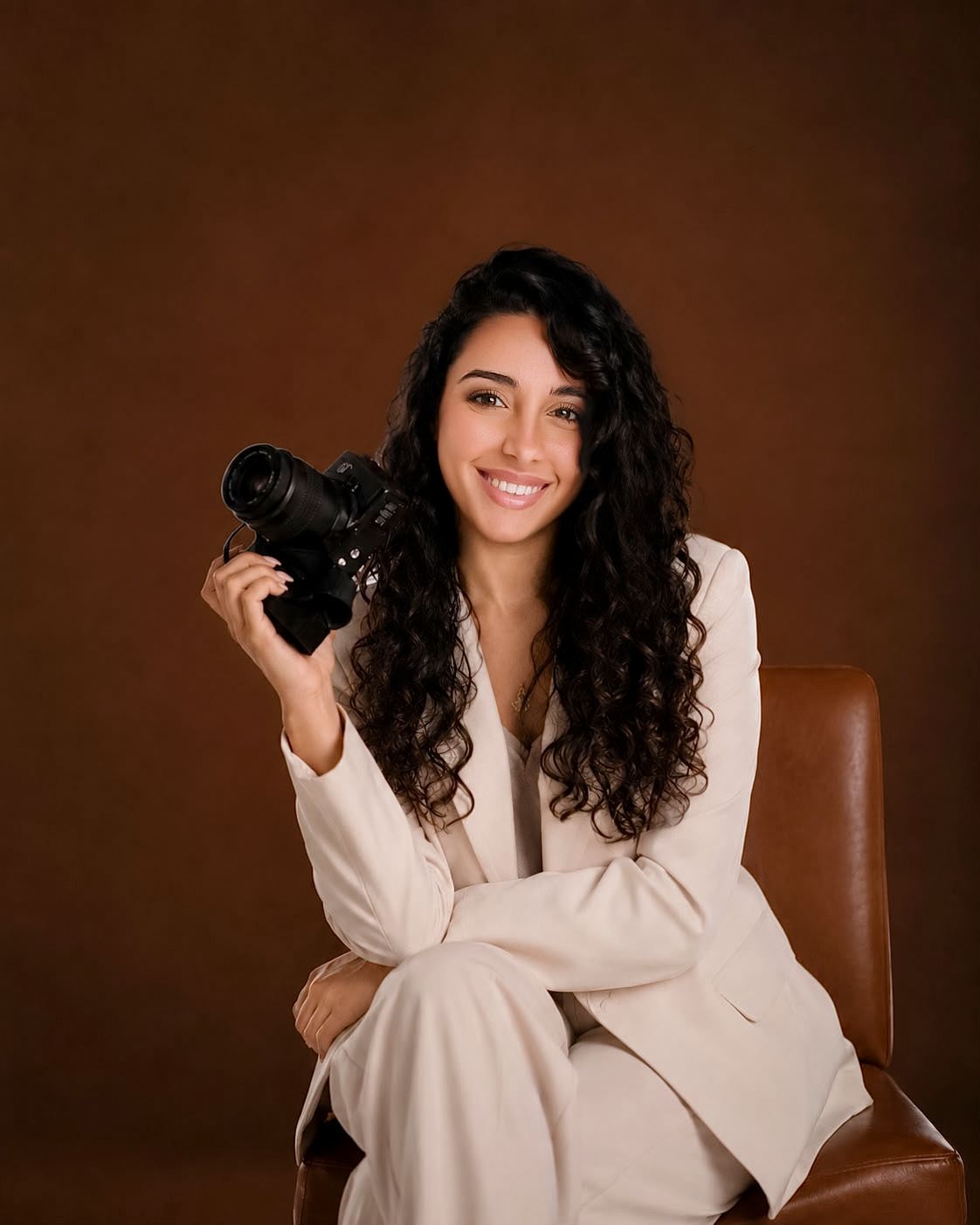 Photographer in beige hat and knit top sits on wicker stool holding professional camera in bright studio setting.