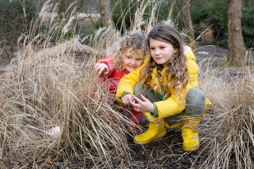 Two young people in bright raincoats exploring tall grasses outdoors on a nature walk.