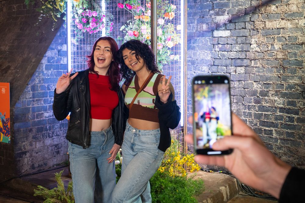Friends pose for a phone photo in front of a neon-lit brick wall with plants and decorative lighting.