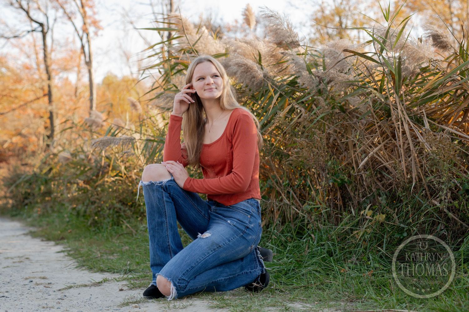 Person in red top and jeans sitting casually on path with fall foliage backdrop.