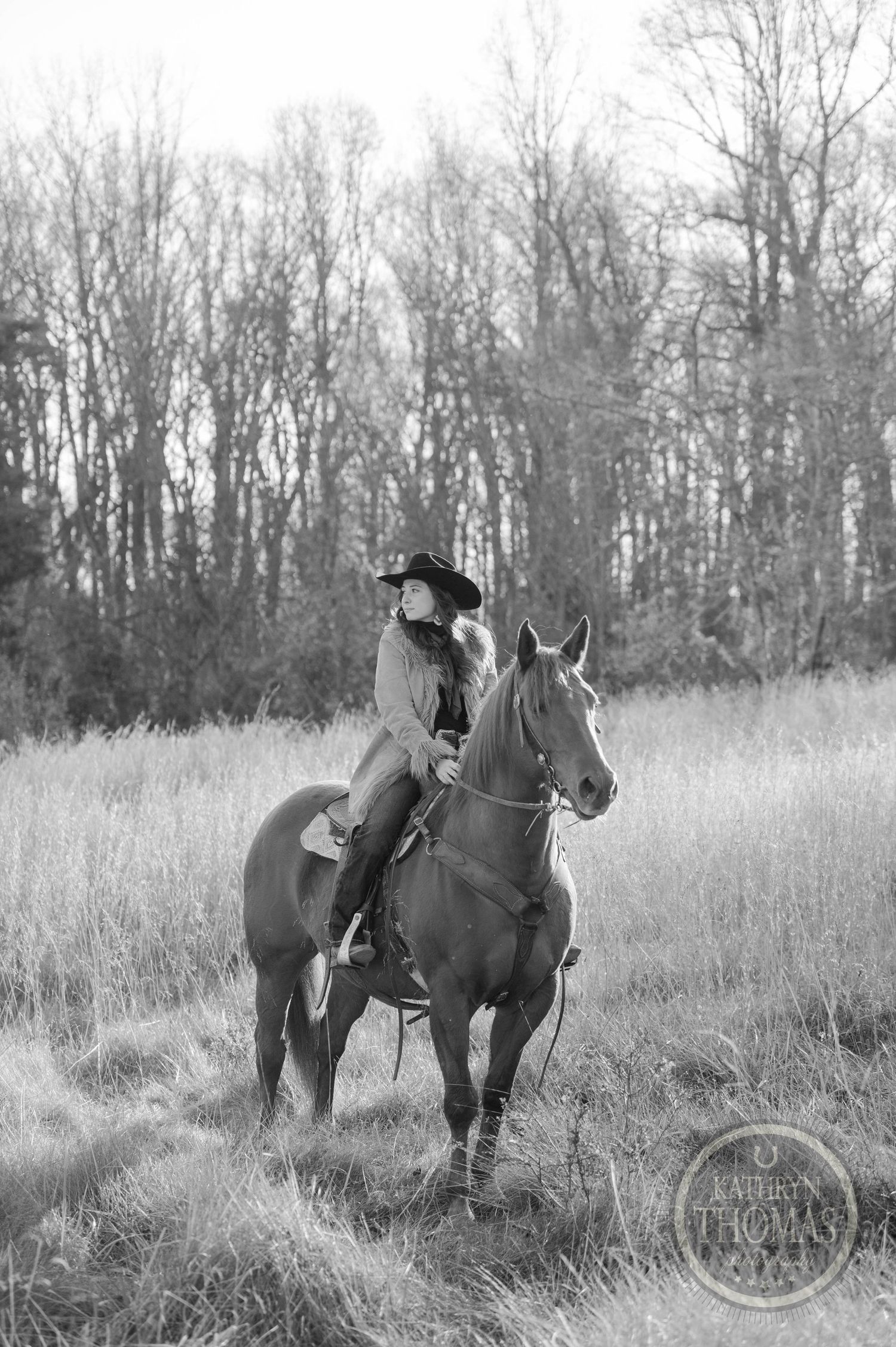 Black and white portrait of an equestrian rider in tall grass with bare trees behind.
