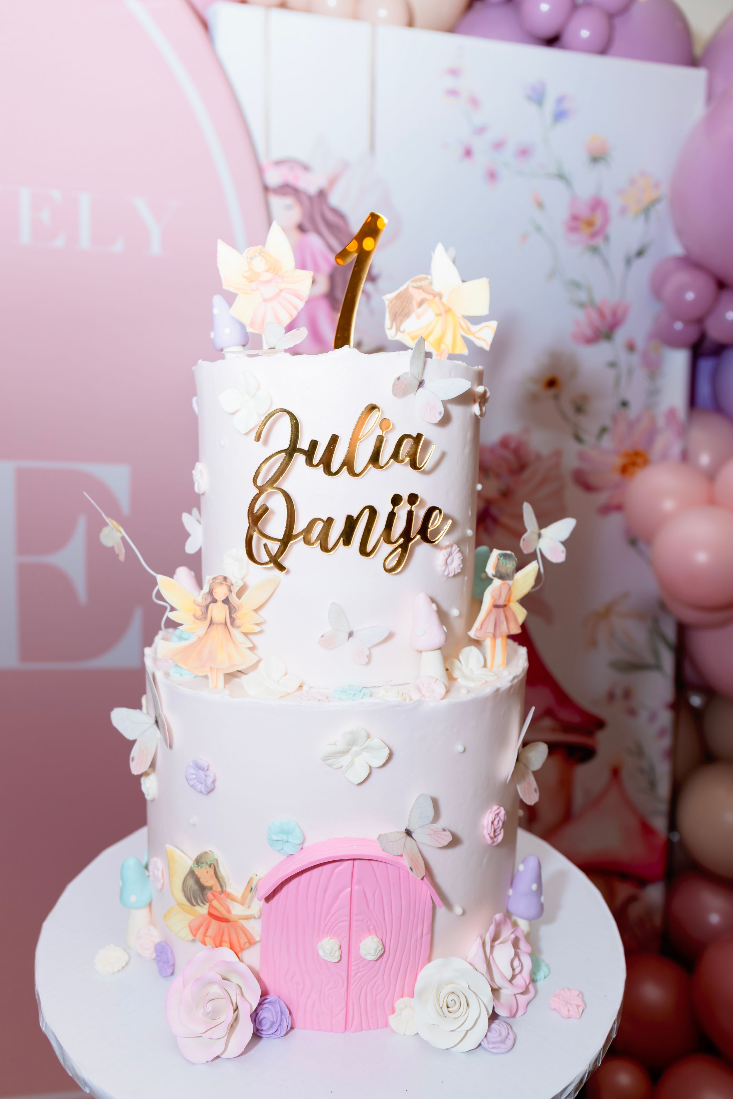 Elegant two-tier fairy themed birthday cake with pink door, floral decorations and gold name topper in pastel colors.