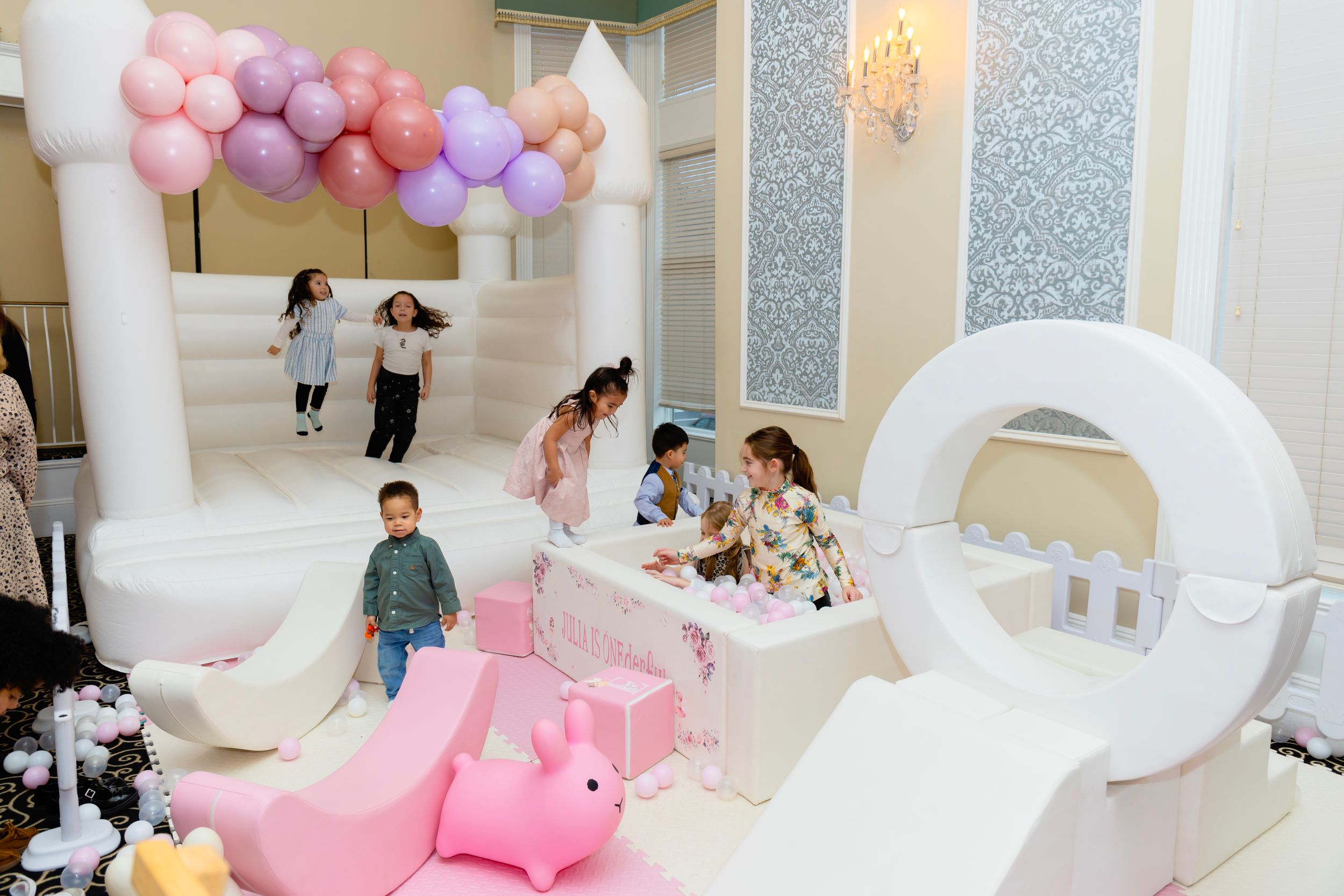 Children playing in a party room decorated with pink and purple balloons, white decor elements, and plush toys.