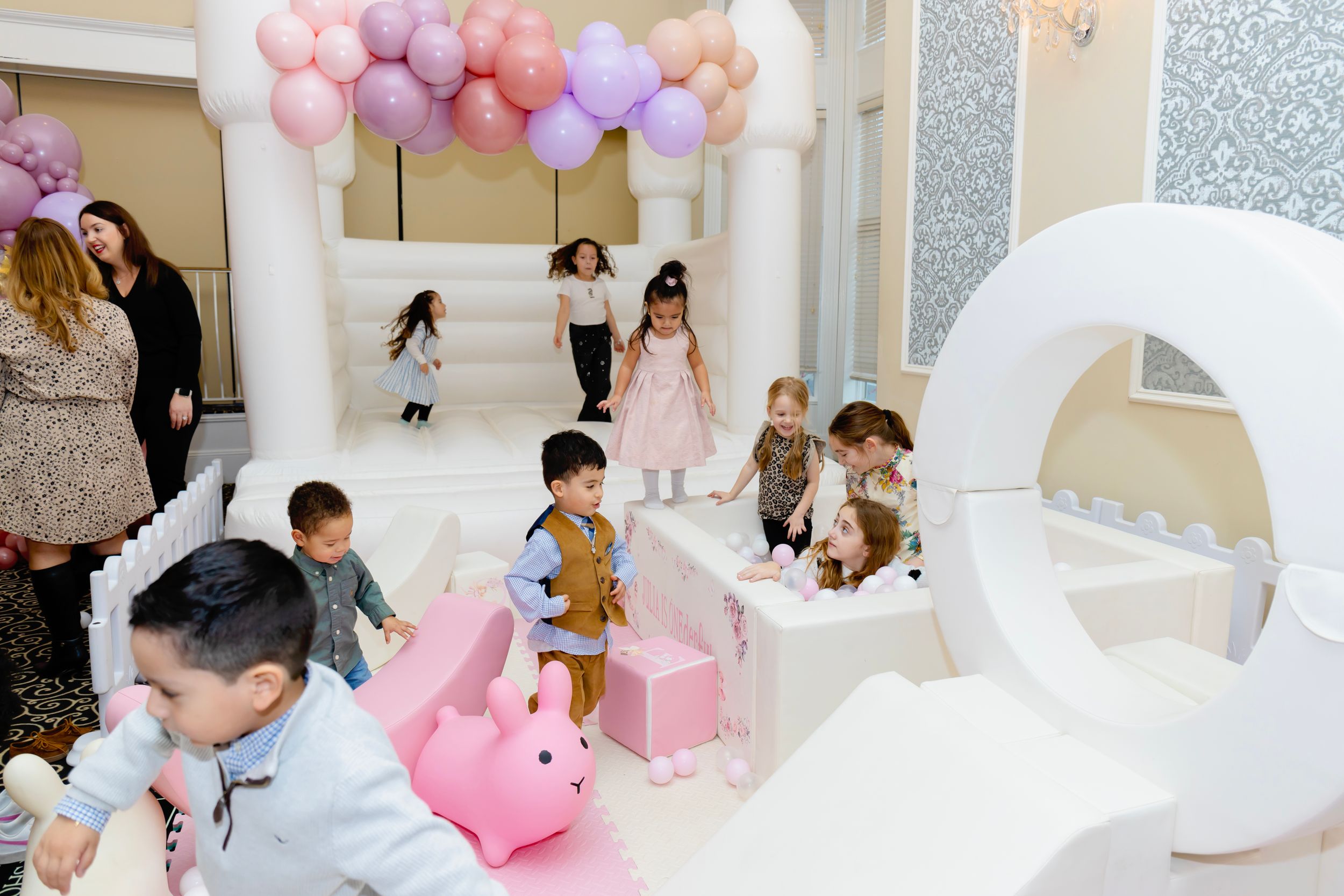 Children playing at a whimsical birthday party with pink balloons, white arches and decorations in a bright indoor space.
