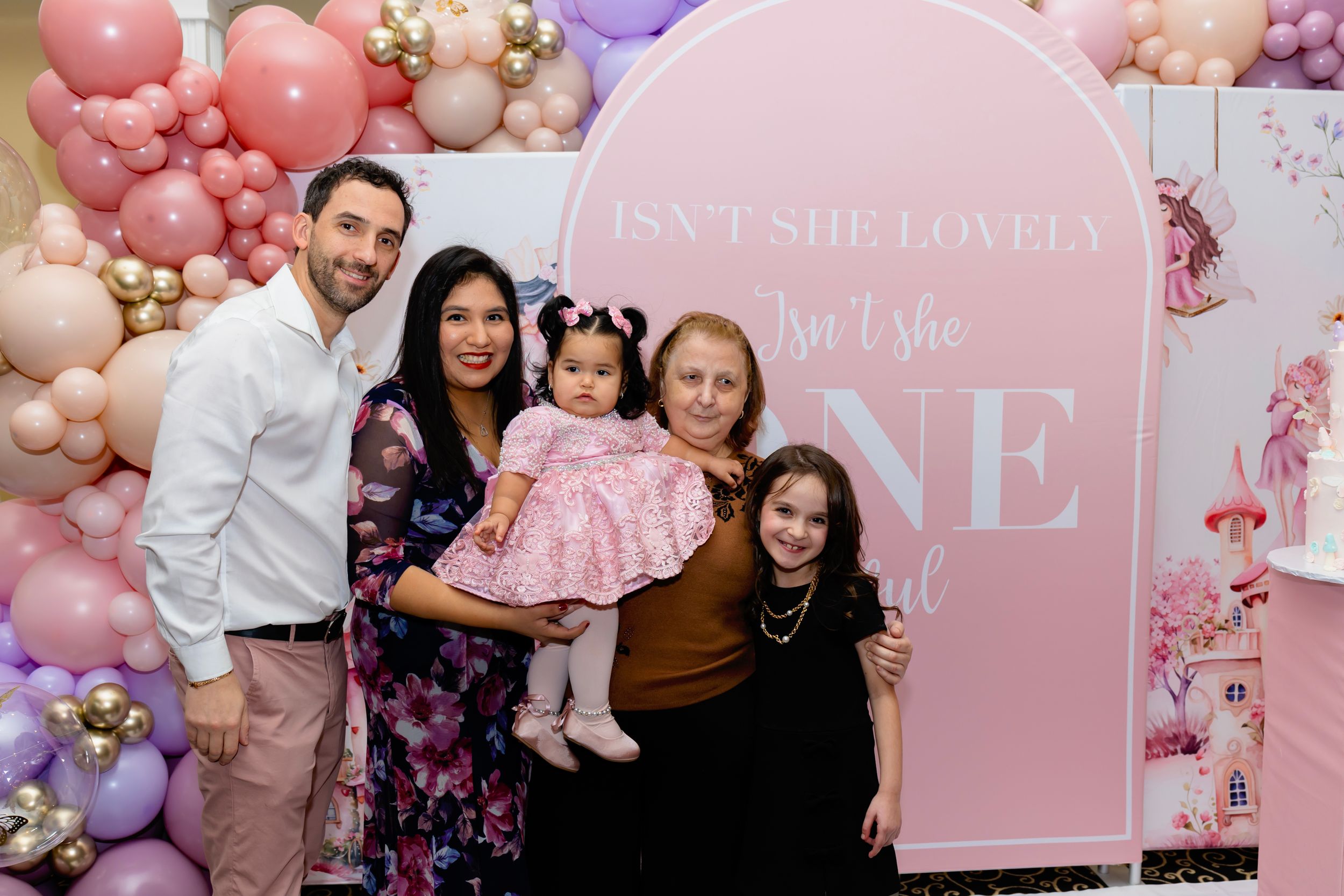 Family group poses together in front of pink balloon arch and decorative backdrop at a first birthday celebration.