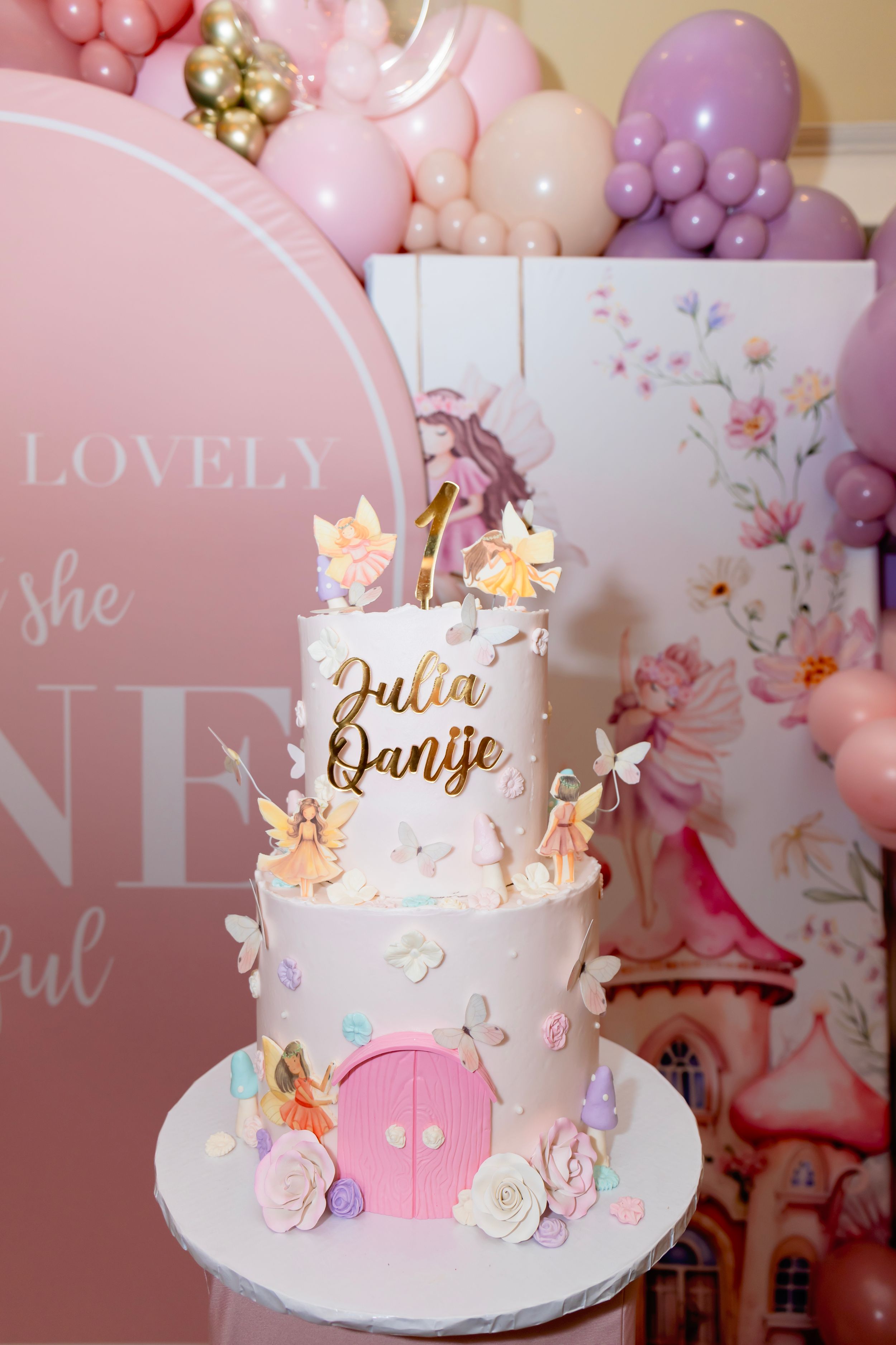 A whimsical pink and white birthday cake with butterflies, flowers and a round door decoration against a balloon backdrop.