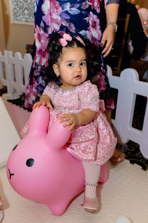 A small child in a pink lace dress sits atop a large pink bunny toy at what appears to be a party venue.