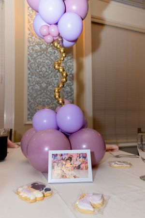 Purple and pink balloon arrangement with decorative gold accents sits on a table with cookies and a small framed photo.