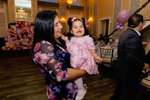 A woman in a floral dress holds a laughing toddler wearing a pink tulle dress at an indoor celebration event.