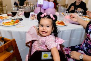 Young child in pink dress smiles from their seat at an elegant party with table settings and pink balloons.